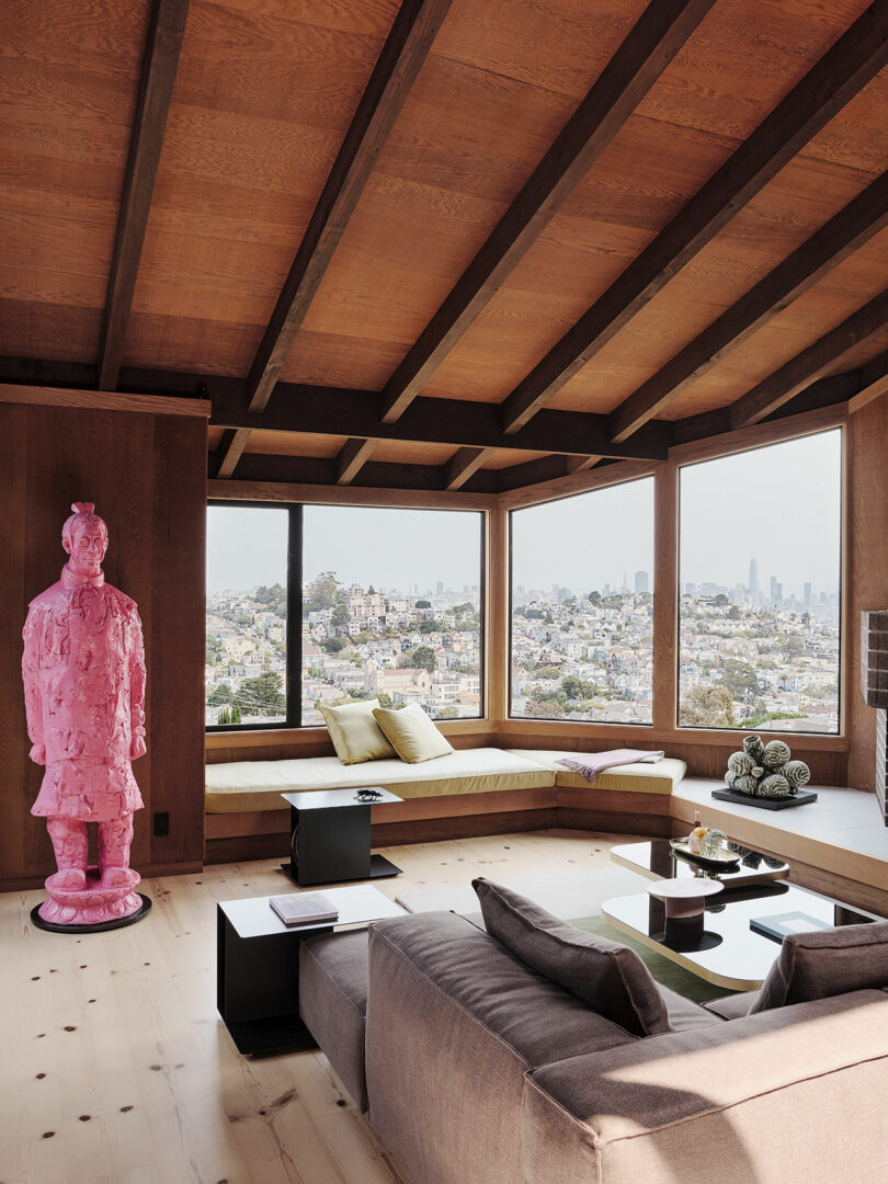 Modern living room with a large window view of a city skyline, built-in seating, neutral furnishings, and a pink statue of a terracotta warrior in the corner.