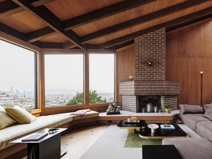 Modern living room with large windows, wood finishes, built-in bench seating, a brick corner fireplace, and a city view in the background.