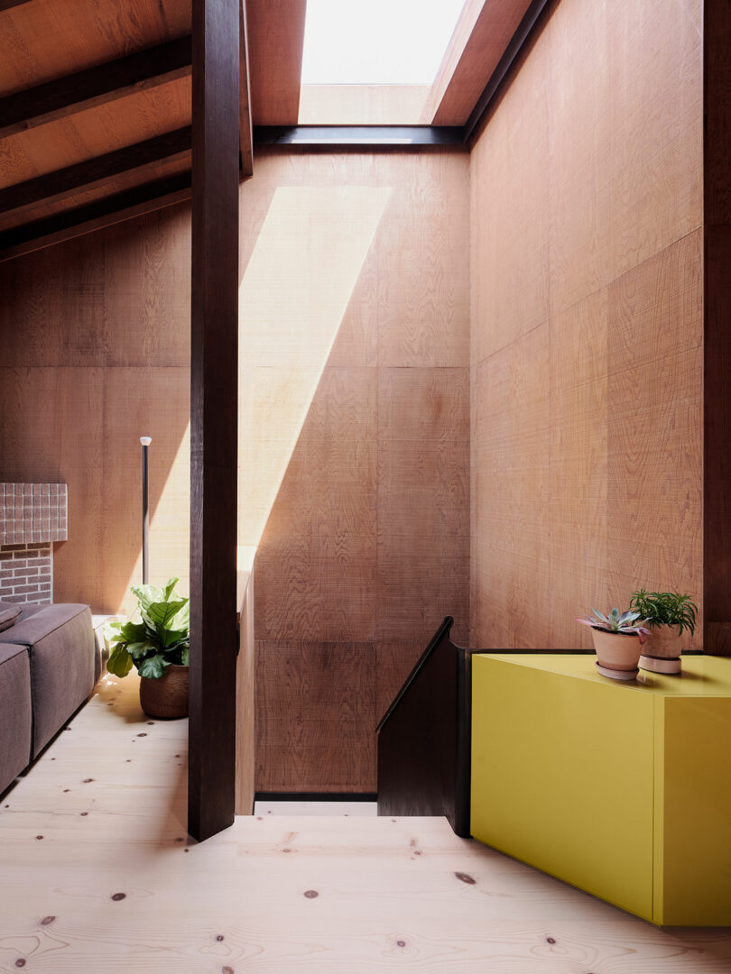 A modern interior staircase with wood-paneled walls, a skylight casting light on the wall, potted plants, and a yellow cabinet beside the stairs.