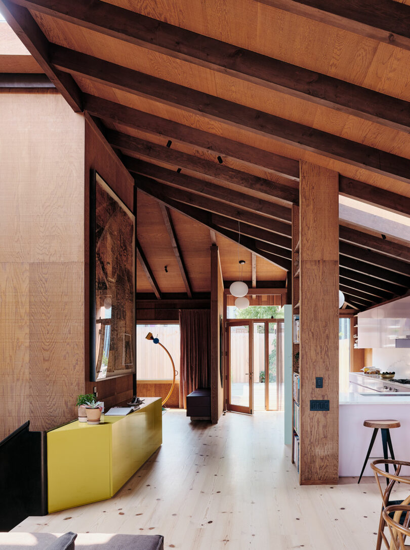 Modern open interior with wood-paneled sloped ceiling, yellow sideboard, light wood floors, and natural light from glass doors leading outside.