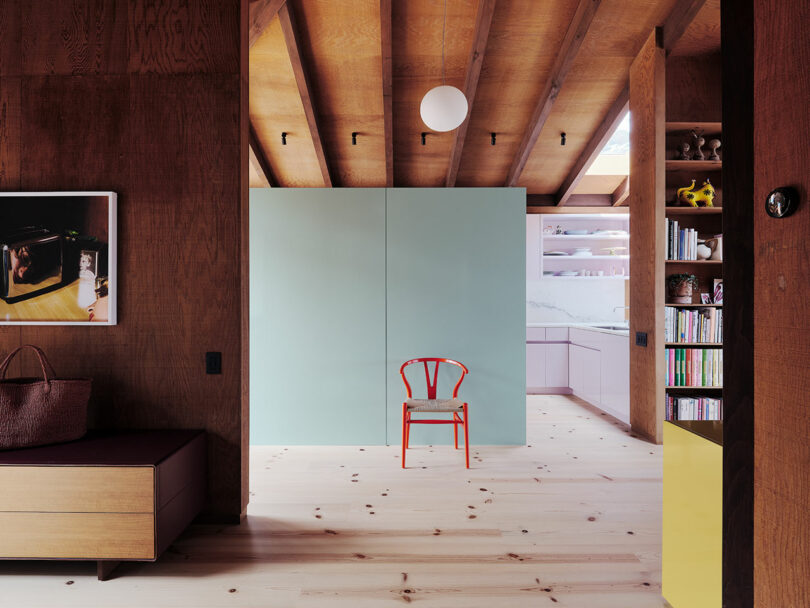 A red chair sits in front of a pale green wall in a modern room with wood beams, open shelving, and a kitchen in the background.