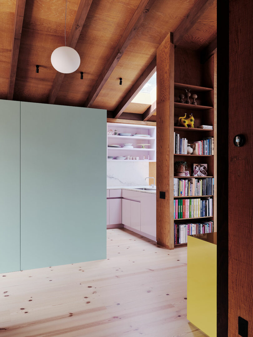 Contemporary kitchen with pastel cabinets, open shelving, and marble backsplash; adjacent bookshelf and a green partition under wooden ceiling beams.
