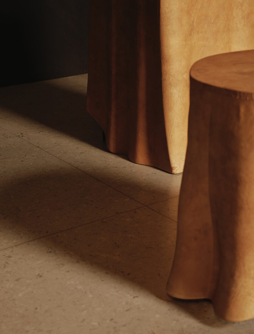 A wooden stool and a wooden table with cloth-like carved drapes stand on a beige stone tile floor, casting soft shadows in warm light.