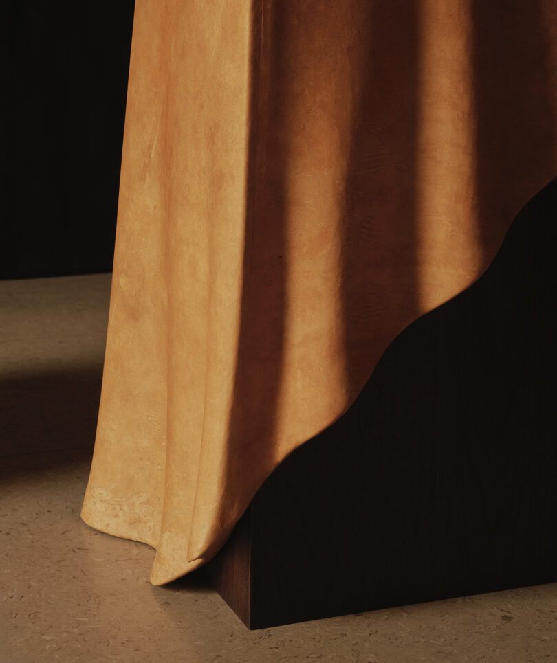 A close-up of a gold fabric draped over the corner of a dark wooden surface, with shadows cast on a beige floor.