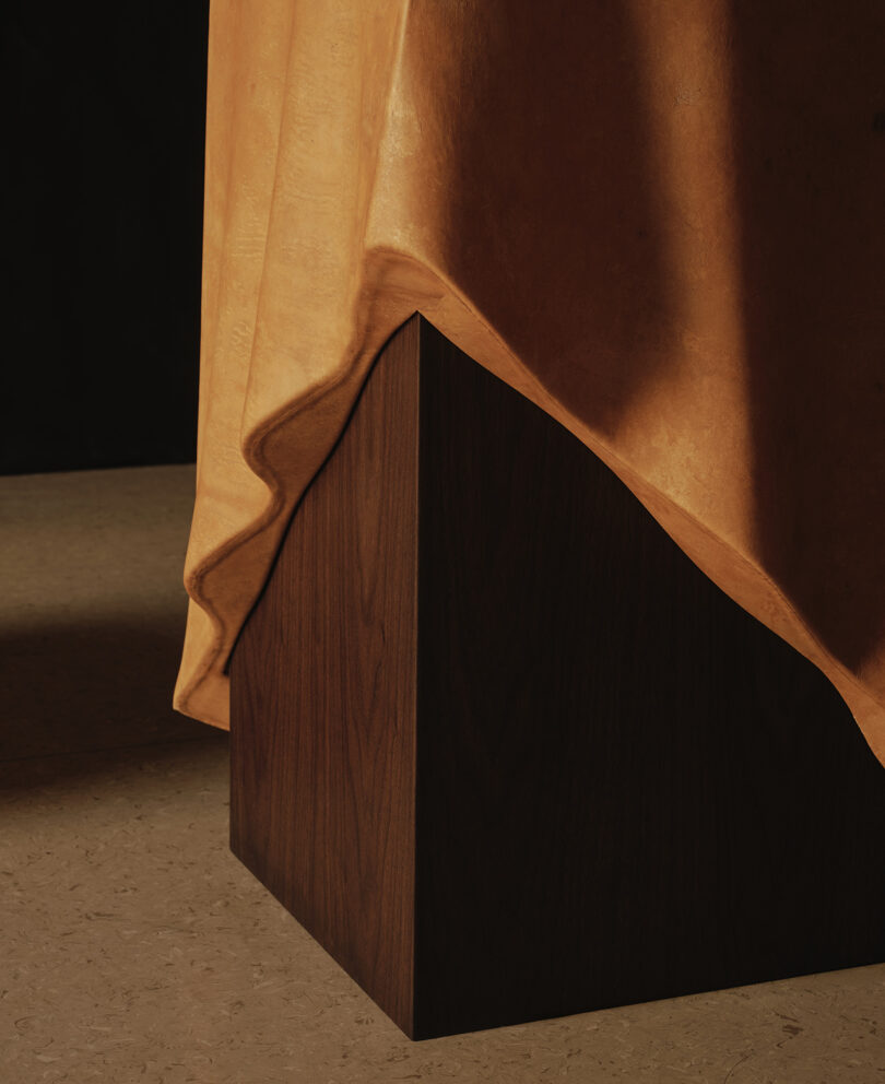 A close-up of a geometric, dark wooden pedestal partially covered by a draped, ochre-colored cloth against a muted background.