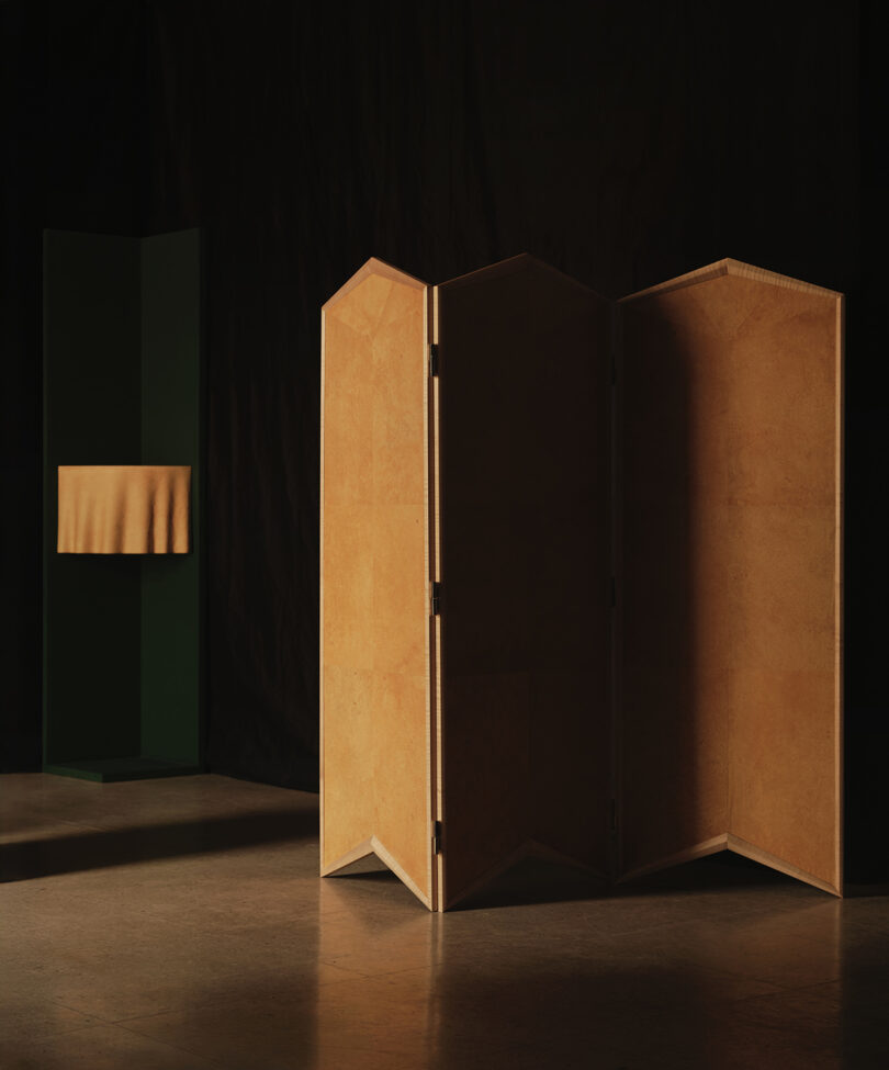 A wooden folding screen stands in front of a dark background, with a green pedestal covered by a beige cloth visible to the left.