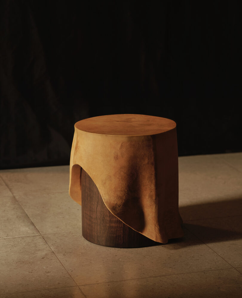 A round wooden stool partially covered with a tan suede-like cloth, placed on a tiled floor against a dark background.