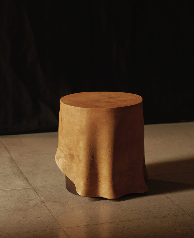 A cylindrical wooden stool covered by a draped, tan leather cloth sits on a tiled floor against a dark background.