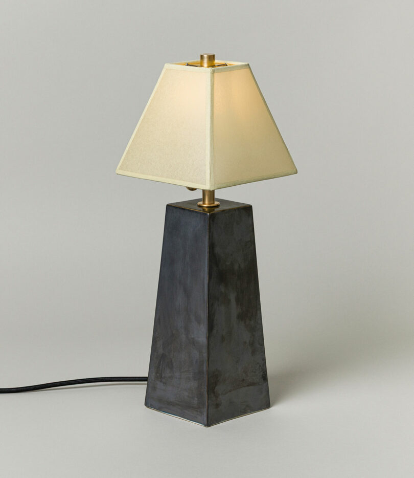 A Virignia Sin lighting piece featuring a black, trapezoidal base and a cream-colored, pyramid-shaped lampshade, illuminated against a plain light background.
