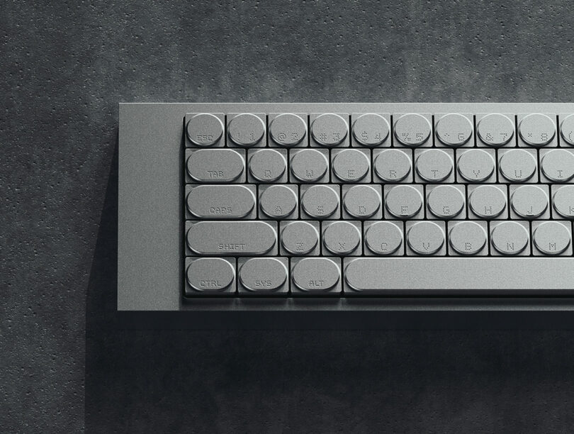 A metallic keyboard with round keys, including labeled buttons such as "CTRL," "SHIFT," and "ALT," rests on a textured gray surface beside a polished cleaver.