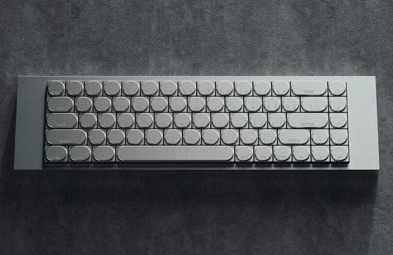 A modern, compact keyboard with round keys sits on a dark, textured surface with horizontal lines and subtle rivet details, resembling the precise design of a cleaver.