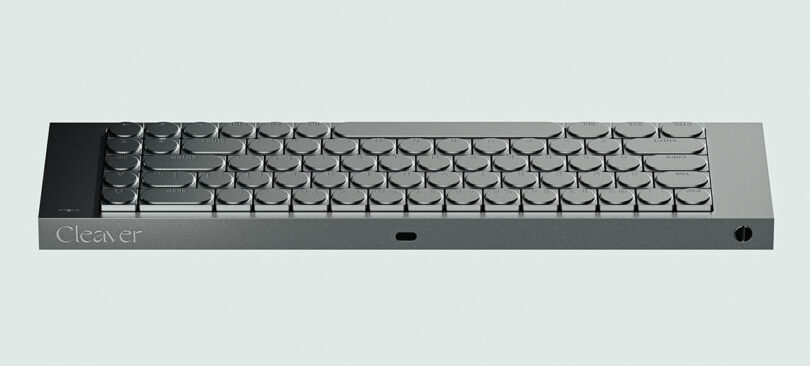 A minimalist, metallic keyboard with round keys arranged in a compact cleaver-inspired layout, viewed from the front against a light background.