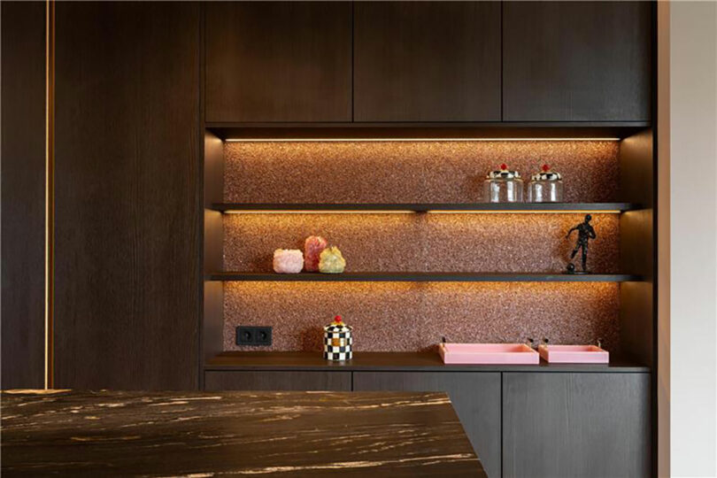 Modern kitchen shelves with brown cabinets, under-shelf lighting, decorative jars, a small statue, pink trays, and a checkered container on a brown textured backsplash.