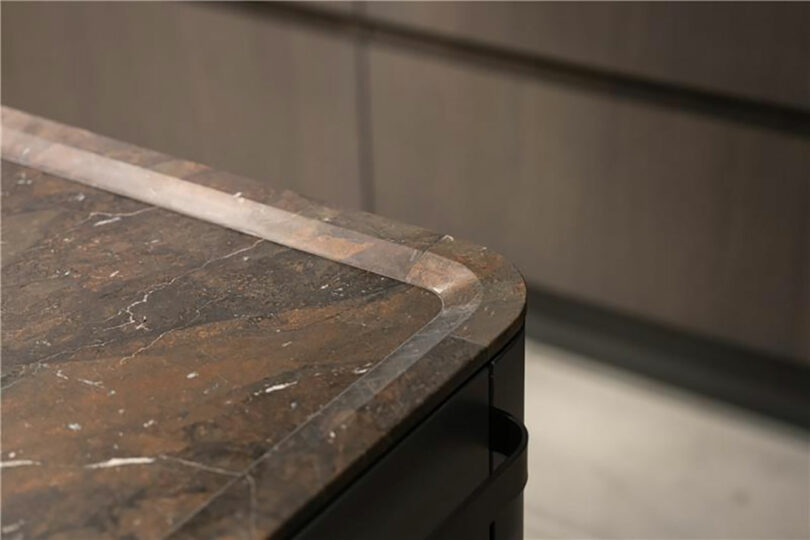 Close-up of a kitchen countertop with a brown marble surface and rounded edges, set against a blurred background of dark cabinetry.