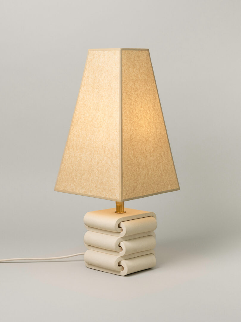A beige table lamp with a rectangular shade and a stacked, sculpted base, reminiscent of Virginia Sin lighting, sits on a plain, light surface