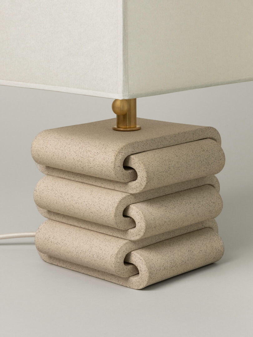 A square lamp with a sculpted, stacked stone base and a white rectangular lampshade, featuring a visible gold fixture and a white power cord—an elegant nod to Virignia Sin lighting design