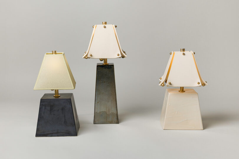 Three square-based table lamps with cream-colored lampshades and gold accents evoke a modern elegance, reminiscent of Virignia Sin lighting, displayed against a plain, light gray background