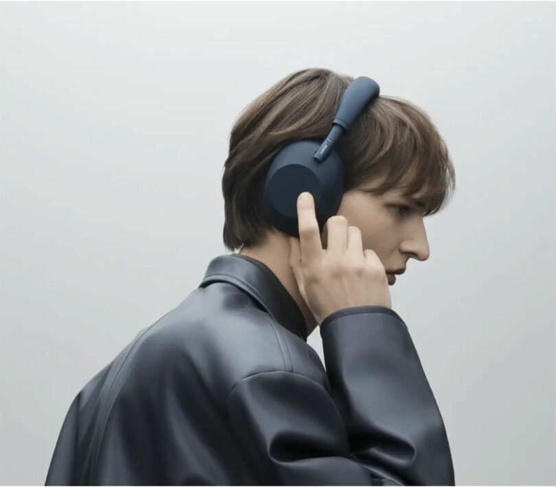 A person wearing a dark leather jacket listens to WH-1000XM6 over-ear headphones, one hand touching the headphones against a plain, light background.