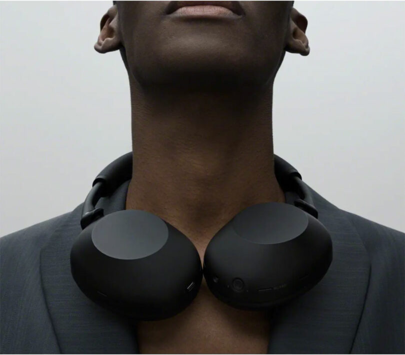 A person wearing a dark blazer has black WH-1000XM6 over-ear headphones resting around their neck against a plain, light background.