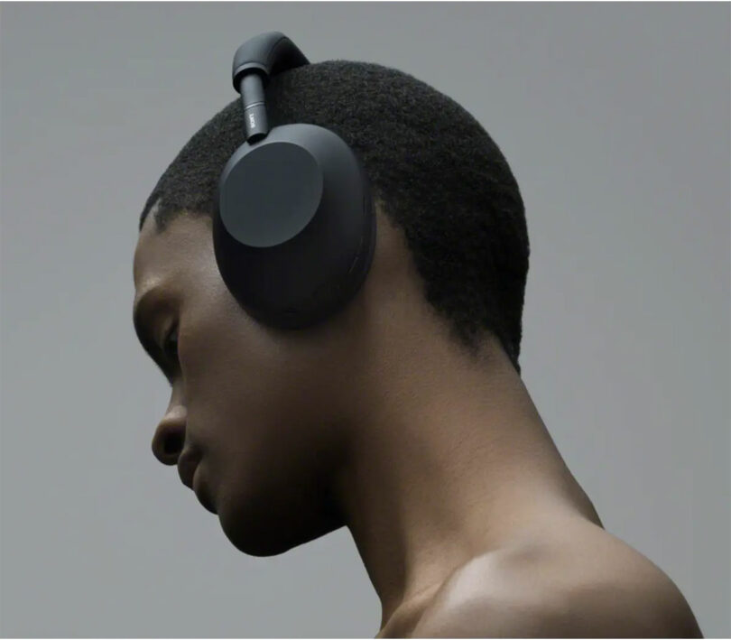 A person with short hair wears large, black WH-1000XM6 over-ear headphones and looks downward against a plain, neutral background.