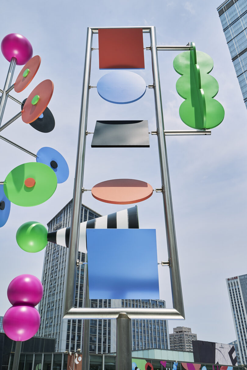 Colorful geometric metal sculptures featuring circles, rectangles, and ovals are displayed outdoors against a backdrop of modern city buildings and a clear sky.