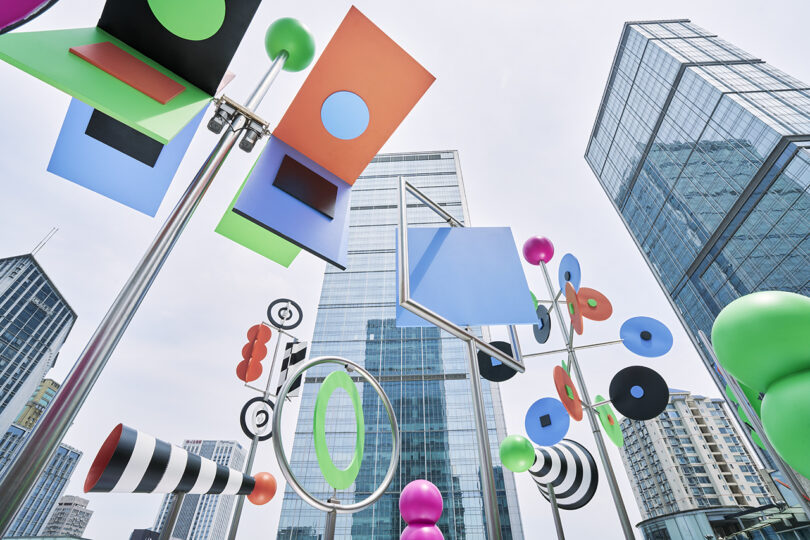 Colorful geometric sculptures are displayed outdoors with tall modern glass buildings in the background, photographed from a low angle.