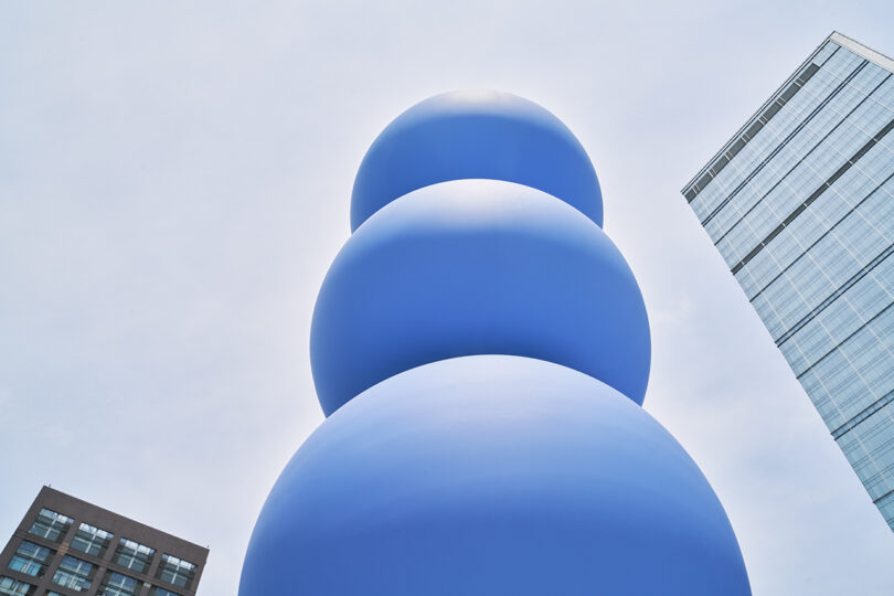 A tall blue sculpture of stacked spheres outdoors, photographed from a low angle, with modern buildings in the background against a pale sky.