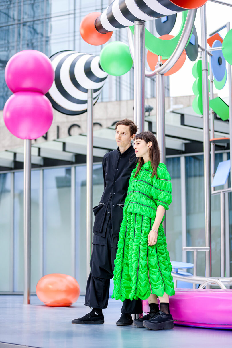 Two people stand next to each other in front of colorful, outdoor abstract sculptures; One bears a black outfit, the other one shiny green dress.