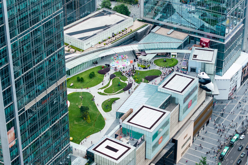 Aerial view of an urban park with winding paths and grass, surrounded by modern glass buildings and busy city streets with pedestrians and vehicles.