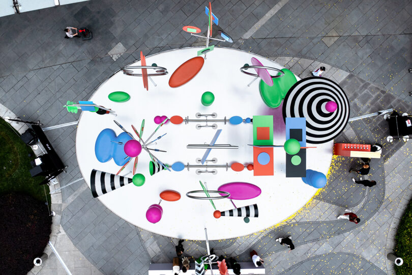 Aerial view of an outdoor art installation featuring colorful geometric shapes, spheres, and black and white striped patterns on an oval white platform with people nearby.