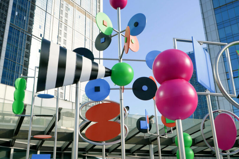 Colorful abstract outdoor sculpture with geometric shapes and spheres invites you to stop, listen, and look, set against modern glass buildings and a clear sky.
