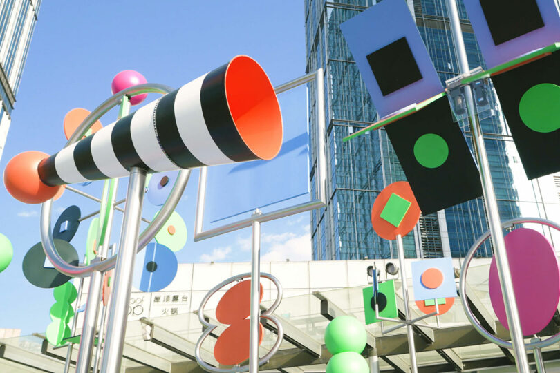 Colorful abstract sculptures with geometric shapes and patterns invite you to stop, listen, look, and enjoy their presence outdoors in an urban area, modern buildings rising gracefully in the background.