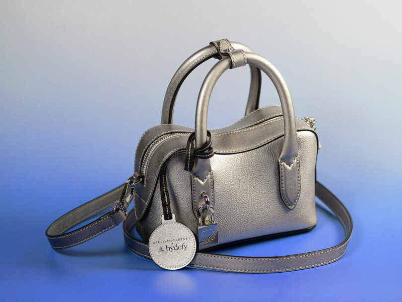 A small metallic silver handbag with two handles, a shoulder strap, zipper closure, and a circular tag, set against a gradient blue background.