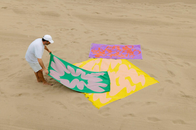 A person on a sandy beach lays down a green and pink patterned towel near two other colorful patterned towels, one yellow and one purple.