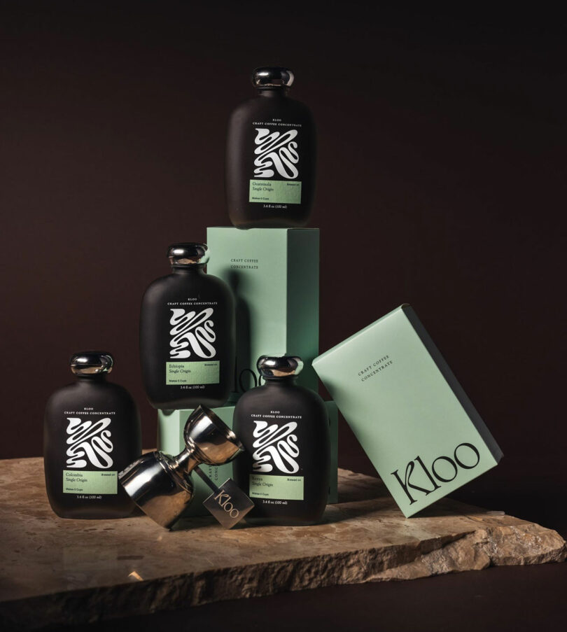 Five black bottles with silver caps and green labels are arranged on a stone surface, alongside green K100 branded boxes, against a dark background.