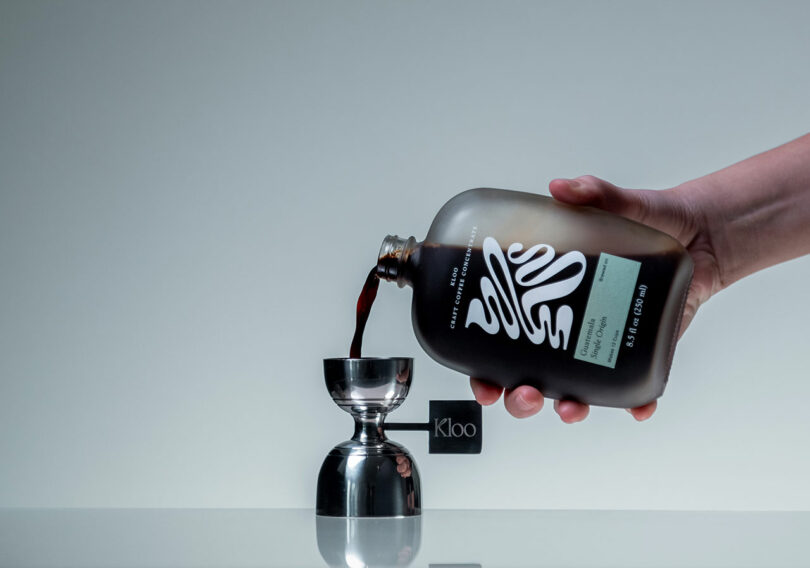 A hand pours dark liquid from a labeled bottle into a metal jigger, with a small black Kloo card visible in the background.