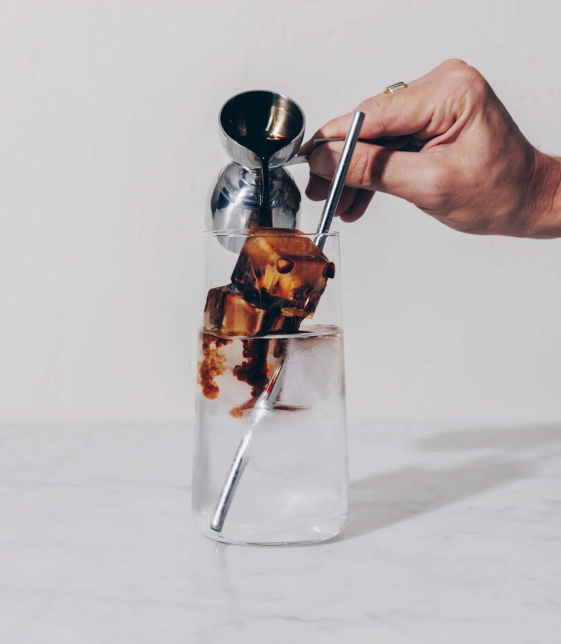 A hand pours liquid from a jigger over large coffee ice cubes in a glass of clear water with a metal straw.