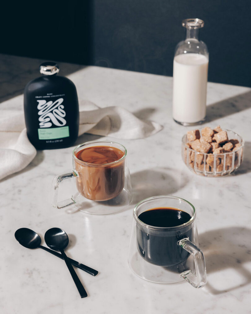 Two glass mugs, one with black coffee and one with coffee and milk, sit on a marble table with a bottle of milk, a bottle of coffee concentrate, sugar cubes, and two black spoons nearby.