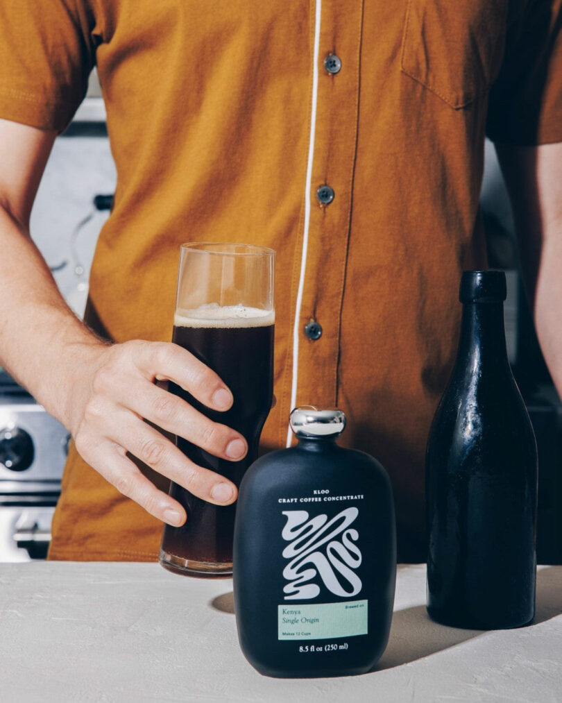 A person in a brown shirt holds a tall glass of dark beverage next to a black bottle and a black bottle labeled "Craft Coffee Concentrate.