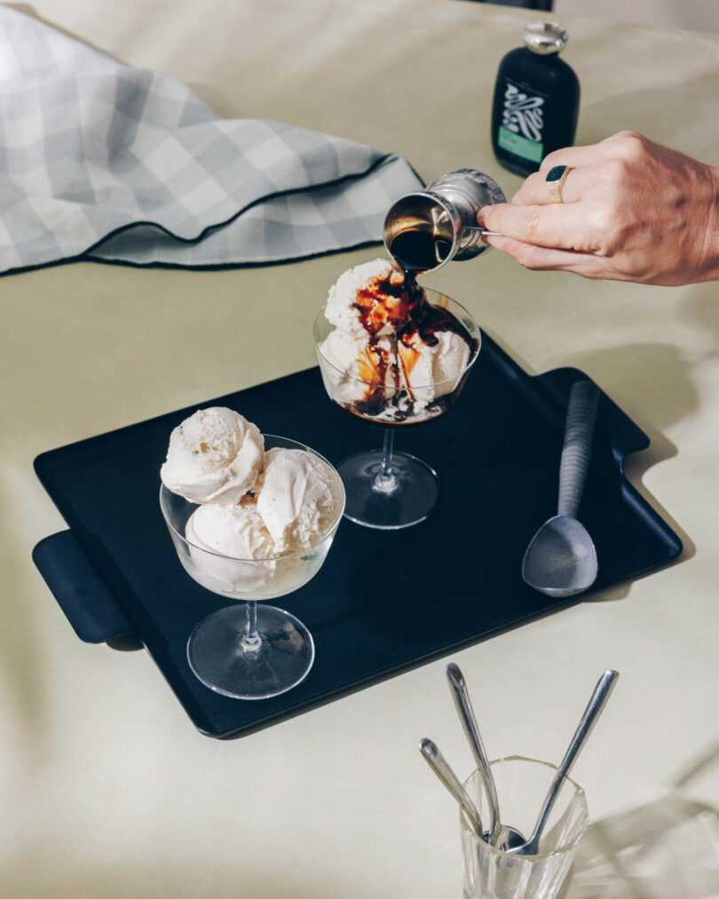 A hand pours espresso over vanilla ice cream in a glass dish, next to another dish with plain ice cream on a black tray, with a scoop and empty glasses nearby.
