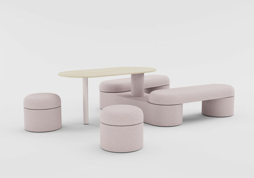 A modern furniture set with a light wood oval table, two cylindrical stools, a backless bench, and a curved seat, all upholstered in light pink fabric, on a white background.