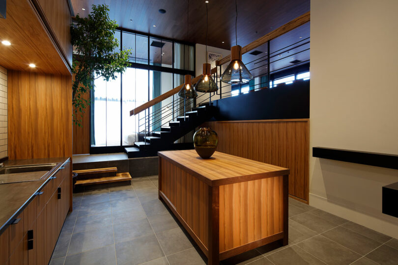 The modern kitchen area in a room from Trunk (hotel) Cat Street, with wooden panel walls and an island, gray tiled floor, elegant black countertop, large windows and a staircase with metal railing on the background