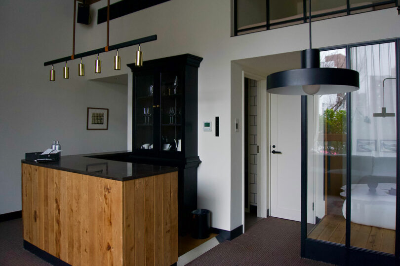 Modern kitchenette/bar at TRUNK(HOTEL) CAT STREET, featuring a wooden island, a sleek black cabinet, hanging lights, and a glass door leading to the bedroom in a minimalist room.