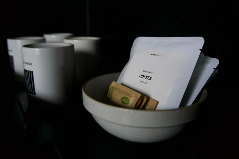 A white bowl holds coffee drip bags and sugar packets next to four white mugs on a dark surface