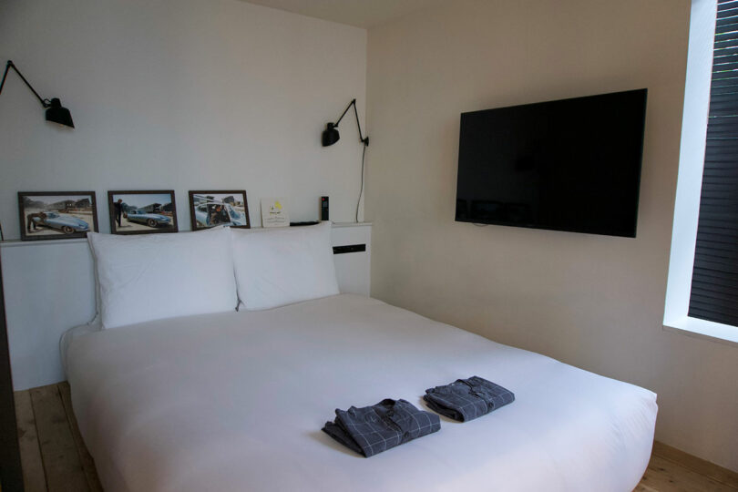A neatly made bed with white sheets with two folded shirts on top, a large wall-mounted TV, framed photos, and two wall lamps in a minimalist TRUNK(HOTEL) CAT STREET bedroom.