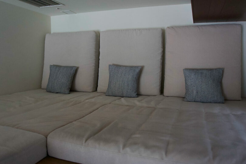 A large beige cushioned seating area with three upright backrests, each featuring a small gray pillow, sits against a white wall