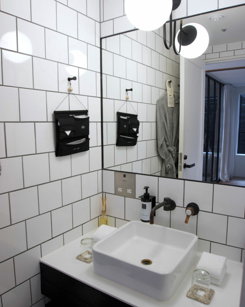 Modern bathroom at TRUNK(HOTEL) CAT STREET with white tiled walls, a rectangular sink, black fixtures, hand soap, two candles, neatly folded towels, and a hanging organizer
