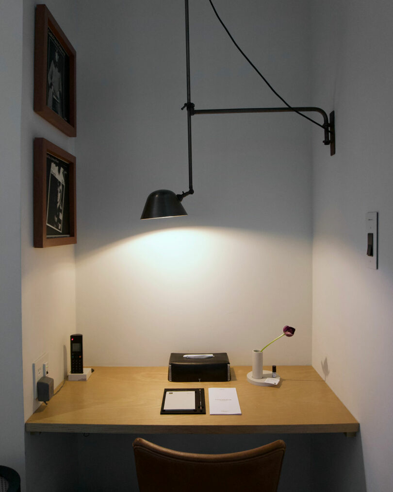 Minimalist desk setup at TRUNK(HOTEL) CAT STREET, featuring a wall-mounted lamp, framed photos, a phone, notepad, papers, tissue box, and a small flower vase against a white wall.