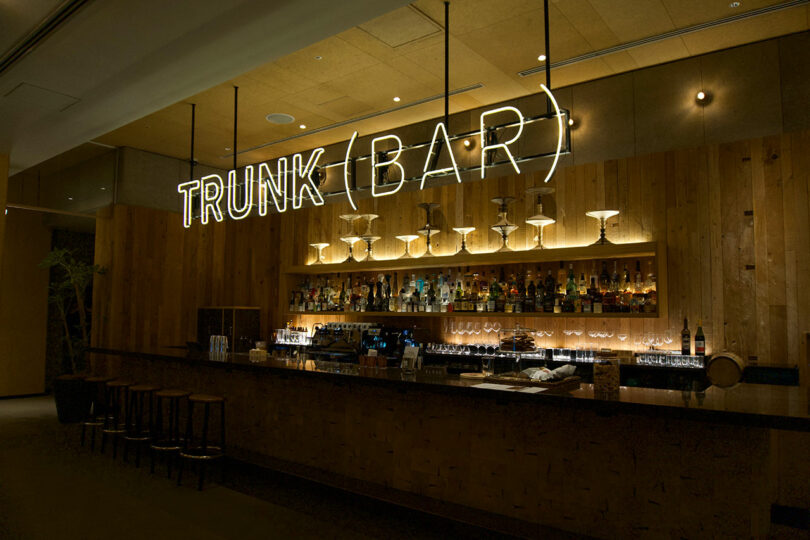 A modern bar with a wooden counter, glassware, and liquor bottles at TRUNK(HOTEL) CAT STREET, featuring a large neon sign reading "TRUNK (BAR)" above the counter.