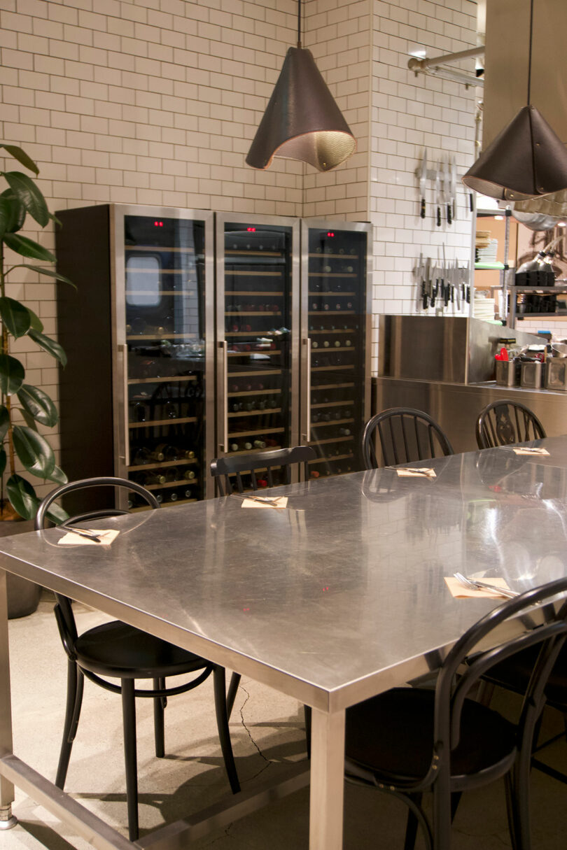 A stainless steel table with black chairs is set next to the kitchen in TRUNK(HOTEL) CAT STREET, featuring wine fridges, hanging lights, and utensils on a tiled wall.